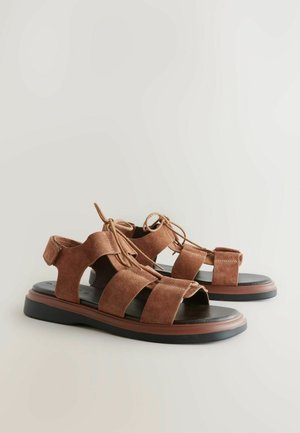 I sandali in suede marrone presentano più fasce larghe, un design con punta aperta e dettagli con lacci. La suola è nera con un accento marrone a contrasto.