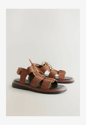 A barna velúr szandálok több széles pánttal rendelkeznek, nyitott orrú kialakításúak és fűzős részletekkel bírnak. A talp fekete, kontrasztáló barna díszítéssel.