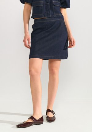 Femme portant une jupe en jean foncé avec une veste courte assortie et des chaussures Mary Jane marron foncé brillantes, debout devant un fond uni.