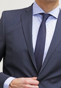 Dunkelblaue Sakkokonfektion mit einer taillierten Passform, kombiniert mit einem hellblauen Hemd, das ein dezentes Polka-Dot-Muster aufweist, und einer dunklen Krawatte.
