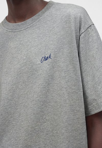 Gros plan d'une personne portant un t-shirt gris avec « Clared » brodé en petits caractères bleus sur le côté gauche de la poitrine.