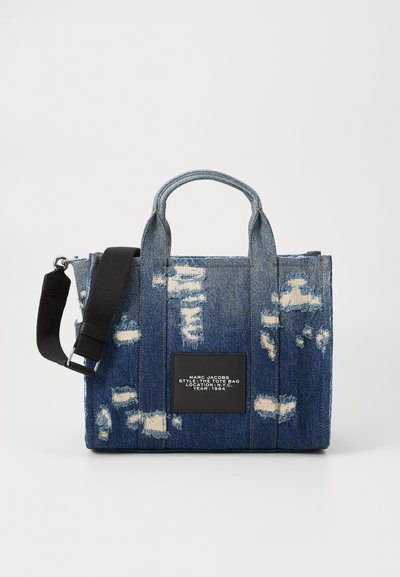 Borsa a tracolla in denim con toppe usurate, manici superiori e una tracolla rimovibile. Presenta un'etichetta nera con testo bianco sul davanti.