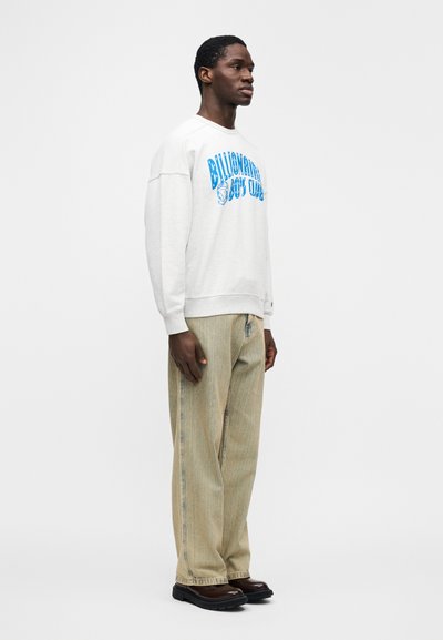 Ανοιχτό γκρι φούτερ με μπλε γραφικό κείμενο, συνδυασμένο με υφασμένα μπεζ παντελόνια και καφέ παπούτσια, παρουσιασμένο πάνω σε απλό φόντο.