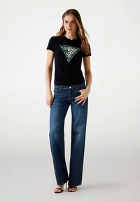 Sort bomulds t-shirt med trekantet turkis og leopardmønster, kombineret med blå denim jeans med lige ben; model iført sorte hæle.