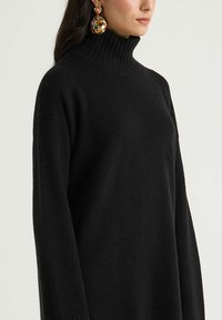 Pull à col roulé noir en maille douce, avec une coupe décontractée, un col côtelé et des manches longues, assorti de grandes boucles d'oreilles décoratives.