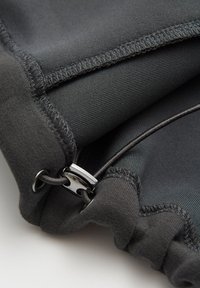Schwarzer Stoff mit glatter Textur, ausgestattet mit elastischem Einfass und einem Metallversteller, der die funktionalen Designdetails des Kleidungsstücks hervorhebt.
