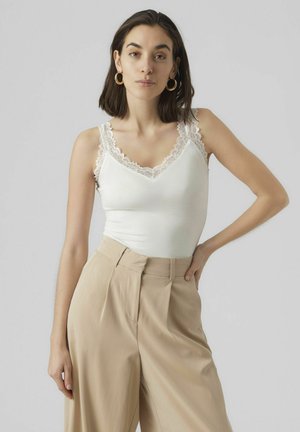 White tank top with lace trim at the neckline and shoulders, paired with high-waisted beige trousers featuring pleated fronts.