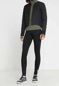 Černá prošívaná bunda přes zelený vysoký krk, doplněná černými legínami a černými teniskami se zlatým akcentem.