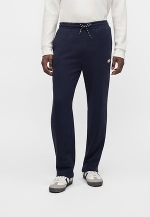 Homme portant un pantalon de survêtement bleu marine avec cordon à la taille et un haut blanc à manches longues, assorti à des baskets blanches et grises sur fond uni.