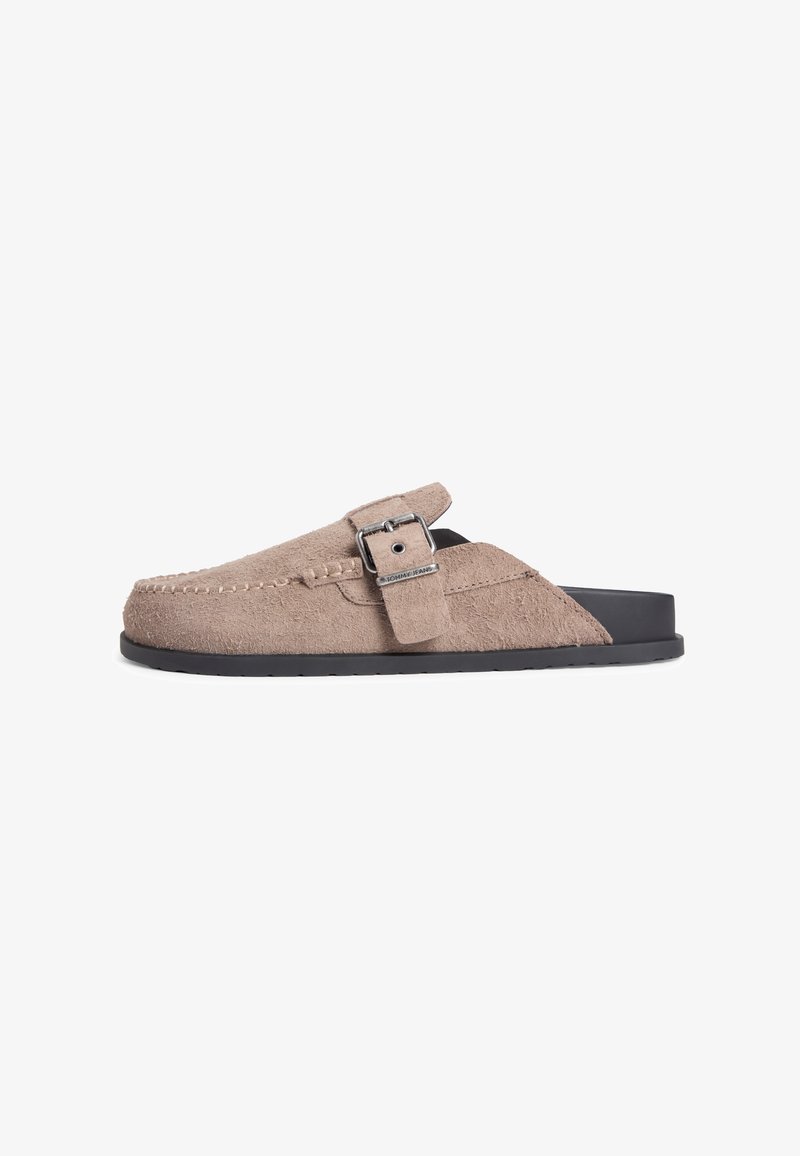 Mule en daim beige avec semelle noire, bride latérale à boucle et détails de couture le long du bord supérieur.