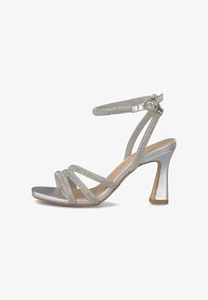 Sandalia plateada de tacón alto con tiras brillantes, hebilla en el tobillo, punta cuadrada y tacón grueso acampanado sobre un fondo blanco.