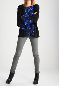 Černý cardigan na modrém vzorovaném topu, šedé úzké kalhoty a černé kotníkové boty. Top má květinové vzory a široké rukávy.