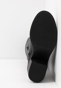 Botín negro con suela texturizada y tacón ancho, posicionado con la suela hacia arriba contra un fondo blanco en la esquina, con la talla marcada como 37.