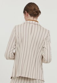 Beige sako s hnědými vertikálními pruhy, jednobarevný design, klopové límce a texturovaná tkanina. Zadní pohled ukazuje přiléhavou siluetu a švy.