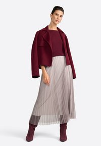 Giacca in misto lana borgogna sopra un top in maglia abbinato, abbinata a una gonna beige fluida e plissettata e stivali al ginocchio in suede borgogna.