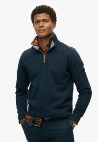 Marineblauer Pullover, mit einem Viertelreißverschluss und grauen Akzenten, über ein kariertes Hemd mit grünen, gelben und roten Farbtönen getragen. Baumwollmischgewebe.