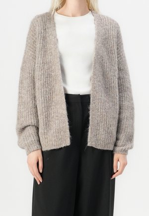 Kobieta ubrana w luźny, teksturowany beżowy kardigan na białym topie i czarne szerokie spodnie, ukazana od ramion do bioder na jednolitym tle.