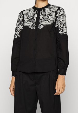 Zwarte blouse met knopen en witte bloemembroderie op de schouders en bovenarmen, gecombineerd met zwarte, loszittende broek.