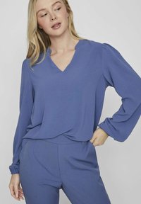 Modra bluzica z dolgimi rokavi in V-izrezom, izdelana iz lahkotnega materiala. Ima nabrano podrobnost na ramenih in sproščen kroj. Nosi se s skladnimi hlačami.