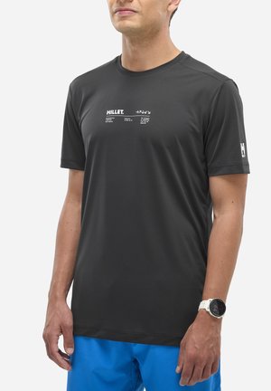 Homme portant un t-shirt MILLET noir à manches courtes avec un logo blanc sur la poitrine et la manche, un short bleu, et une montre connectée blanche au poignet gauche.