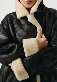 Melna mākslīgā kašmira mētelis ar mīkstu bēšas krāsas apkakli un piedurknēm. Dizainam ir raksturīga teksturēta apdare, brīvs griezums un plata apkakle.