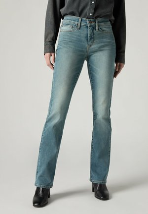 Person trägt hellblaue Jeans mit geradem Bein, schwarze Stiefel mit Absatz und ein dunkles Langarmshirt, die Hände entspannt seitlich.