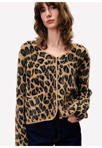 Jonge vrouw met golvend bruin haar, gekleed in een pluizige cardigan met luipaardprint en knopen, met een hand in haar zakken en donkerblauwe spijkerbroek.