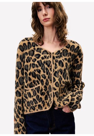 Jeune femme aux cheveux bruns ondulés portant un cardigan en fausse fourrure léopard avec des boutons et un jean bleu marine, main dans la poche.