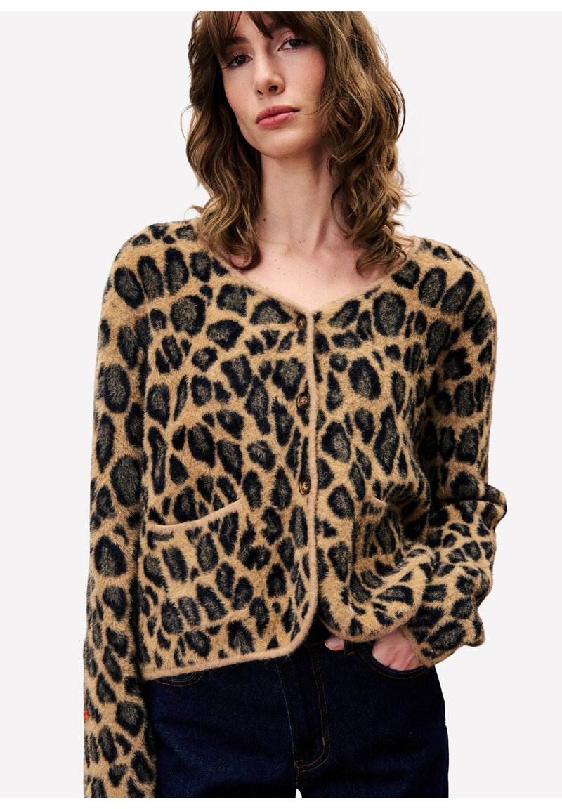 Jonge vrouw met golvend bruin haar, gekleed in een pluizige cardigan met luipaardprint en knopen, met een hand in haar zakken en donkerblauwe spijkerbroek.