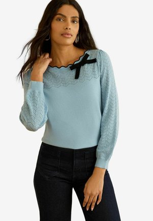 Kobieta w jasnoniebieskim dzianinowym swetrze z koronkowymi rękawami, dekoltem z falbanką i czarną kokardką, w połączeniu z czarnymi spodniami z wysokim stanem.