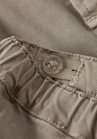 Valgepruun riidekanga lähivõte, millel on sile tekstuur, läbipaistev nööp ja peenelt õmmeldud servad volditud disainiga.