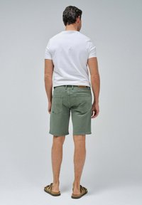 Pantalones cortos de algodón verdes con corte recto, bolsillos traseros y parche de cuero marrón; combinados con una camisa blanca de manga corta y sandalias.