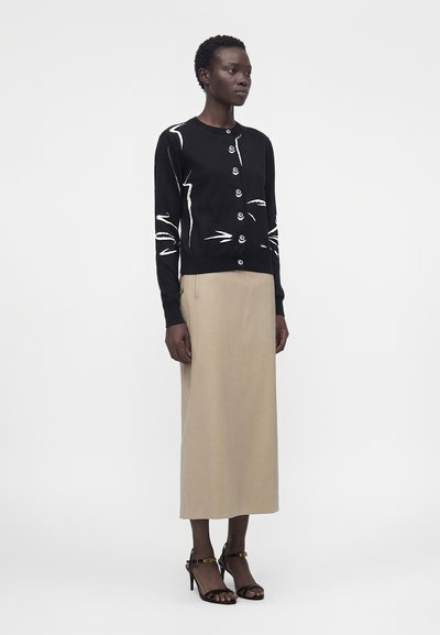 Γυναίκα όρθια, φορώντας μαύρη ζακέτα με κουμπιά και λευκά αφηρημένα σχέδια, μπεζ μίντι φούστα και μαύρα πέδιλα με τακούνια και λουράκια στον αστράγαλο.