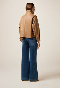 Giubbotto cropped in pelle marrone con colletto alto, abbinato a jeans in denim blu a gamba larga con borchie decorative lungo le cuciture esterne.