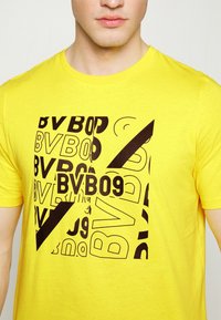 Camiseta de algodón amarilla con un audaz estampado gráfico negro que presenta letras y formas superpuestas, incluyendo "BV09" y un acento de línea diagonal.