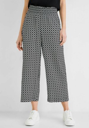 Vrouw die zwart-witte wijde broek met cirkelpatroon en witte sneakers draagt, staand tegen een effen achtergrond.
