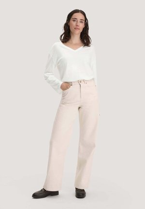 Frau mit dunklen Haaren, die ein weißes langärmliges V-Ausschnitt-Top trägt, hellbeige weite Hosen und schwarze Schuhe, steht mit einer Hand in der Tasche.