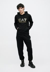 Zwarte hoodie met gouden logo en bijpassende zwarte joggingbroek. De stof lijkt zacht, met een comfortabele pasvorm en elastische manchetten. Zwarte sneakers.