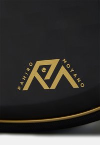 Borsa nera con un logo dorato che presenta forme geometriche e il testo "RAMIRO MOYANO," bordata da una zip dorata e dettagli testurizzati.