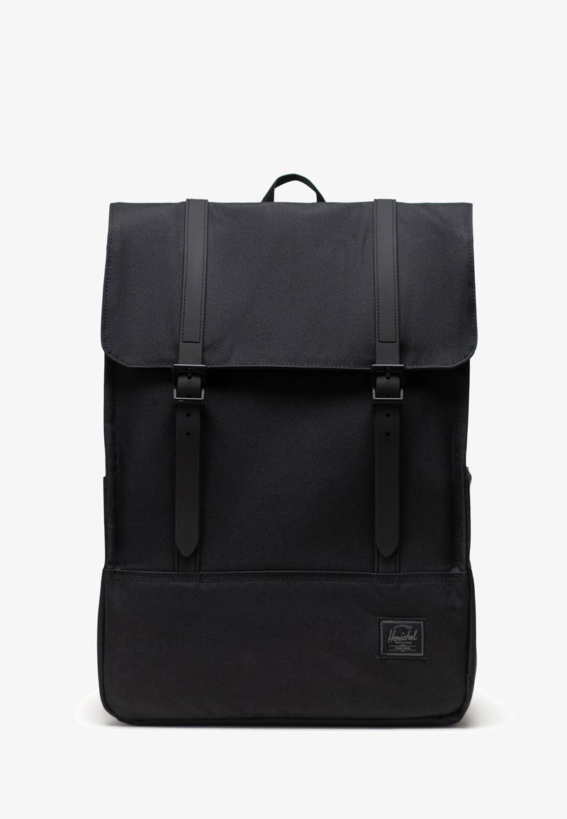 Sac à dos en tissu noir avec un rabat de fermeture, deux bretelles proéminentes, une forme rectangulaire et un écusson logo à l'avant. Surface texturée et design minimaliste.