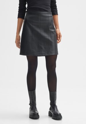 Jupe A-line en cuir noir avec un design en panneaux, présentant une texture lisse. Associée à des bottines noires et des collants opaques.