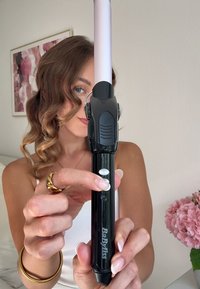 Frau mit lockigem Haar hält und zeigt auf einen schwarzen BaByliss Lockenstab in einem hellen Raum mit rosa Blumen in der Nähe.
