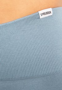 Lichtblauwe ribstof tailleband met een wit label met de tekst "SMILODOX" aan de bovenkant, gestructureerd oppervlak, zacht materiaal.