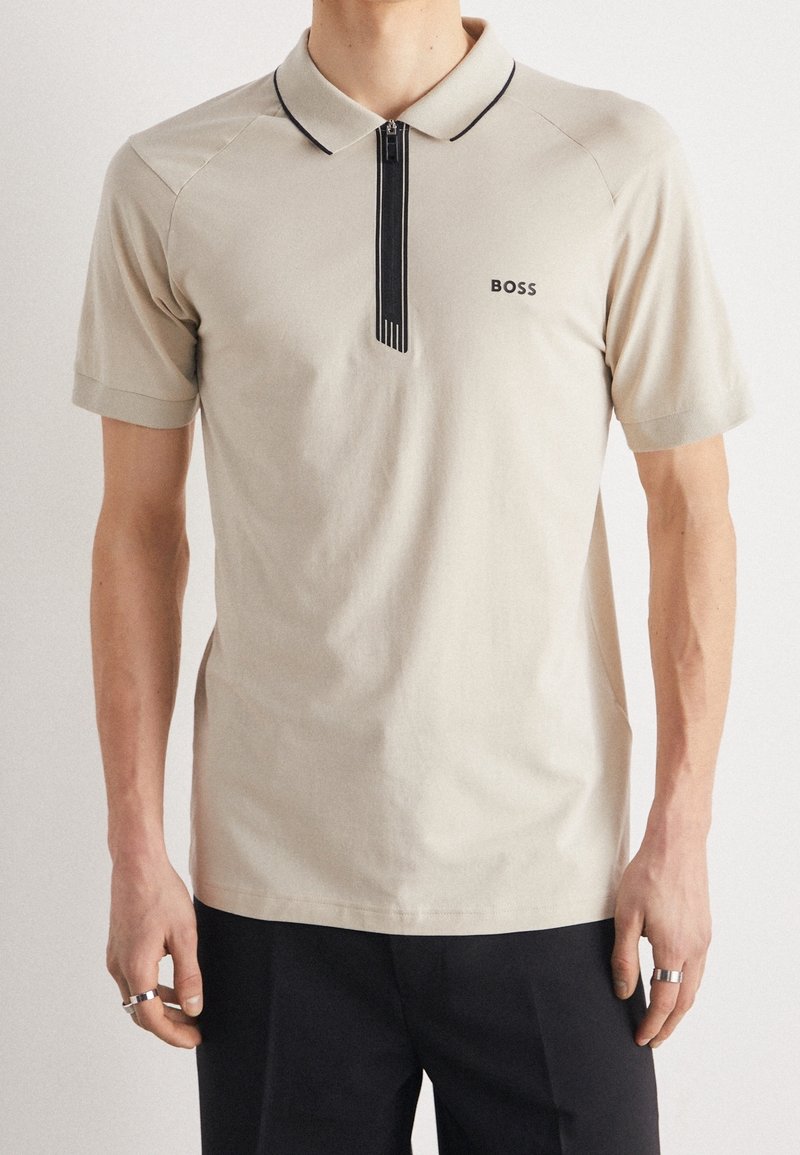 T-shirt polo beige avec un col zippé, un liseré noir et une signalétique discrète. Fabriqué dans un matériau doux, avec des manches courtes et une coupe ajustée.
