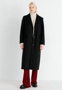 Zwarte lange jas met een gekartelde revers, twee knopen en zijzakken. Gekleed over een crèmekleurige coltrui en vergezeld van bordeauxrode broek met wijde pijpen.