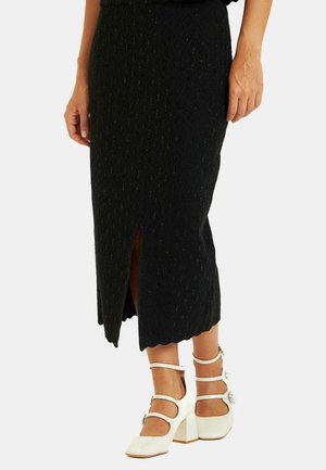 Falda midi de punto negra con un patrón de diamante texturizado, con una abertura en la parte delantera. Combinada con zapatos de tacón bloque blancos con correas.