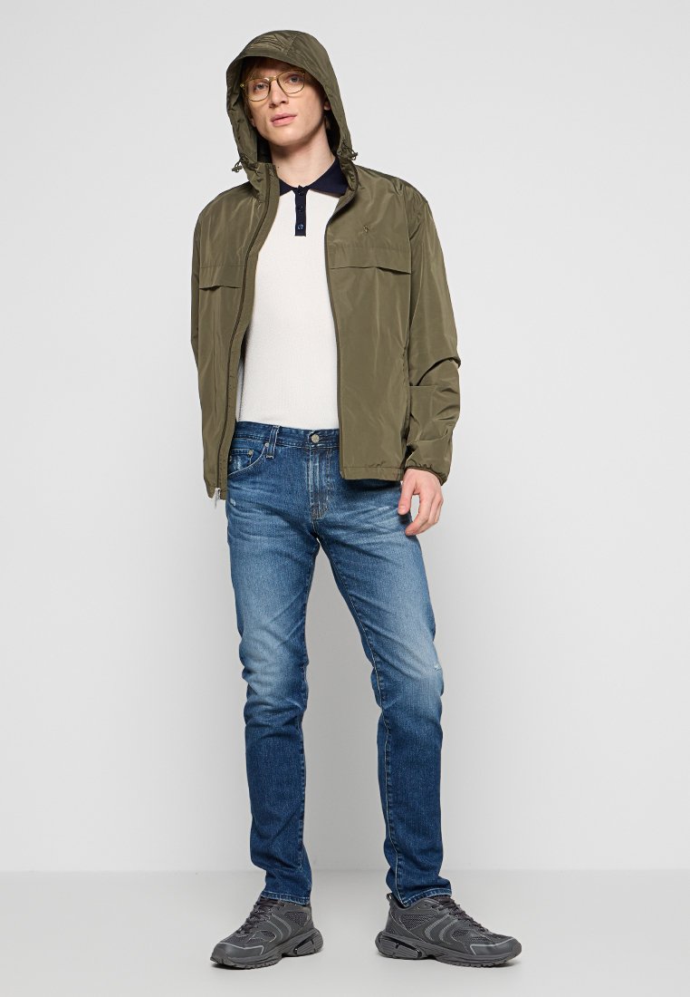 Leichte olivgrüne Kapuzenjacke, weißes Strickoberteil, blaue Jeans und graue Sportschuhe. Zu den Merkmalen gehören doppelte Brusttaschen und ein Kragen.