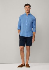 Camicia azzurra chiaro button-up con maniche arrotolate, abbinata a pantaloni corti blu navy e scarpe slip-on marroni. Design semplice e casual con una vestibilità rilassata.