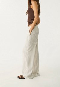 Pantalon léger à rayures et jambes larges en tissu doux ; doté d'une ceinture élastique et de poches latérales. Associé à un débardeur marron et des sandales.