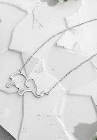 Collana argento a forma di elefante con una coda a forma di cuore, caratterizzata da un design minimalista e una catena delicata, poggiata su una superficie testurizzata.